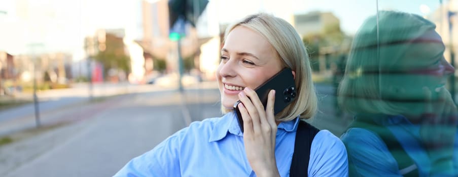 Lifestyle portrait of happy blond woman, chatting over the phone, answers a call, laughing and smiling, standing on street and looking cheerful.