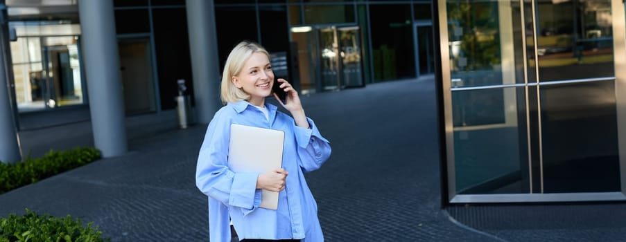 Image of young office woman, employee standing near company building with documents and notebooks, answers phone call, talking on smartphone and smiling, wearing blue collar shirt.