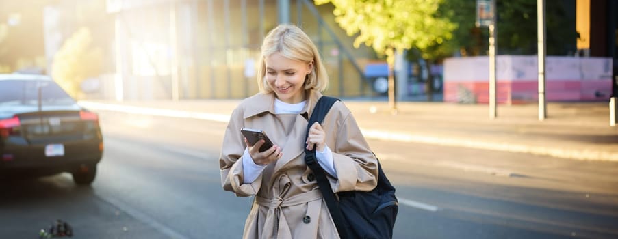 Smiling blonde woman in trench coat, standing on street, using mobile phone app, holding backpack, student with smartphone waiting near road.