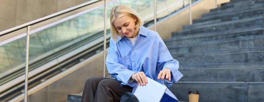 Portrait of young street-style girl, student with backpack, putting away laptop, packing, sitting on stairs with cup of coffee, wearing blue shirt.