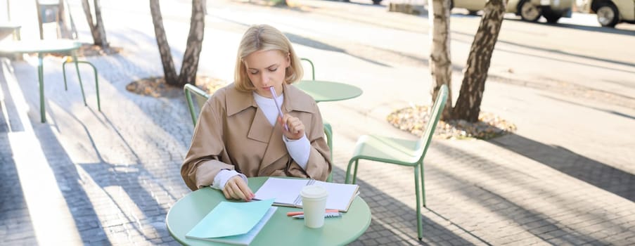 Portrait of young woman thinking while doing homework, sitting in outdoor cafe alone, drinking coffee and writing in notebook, looking thoughtful, pondering smth.