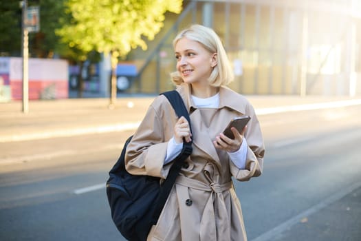 Portrait of young blonde woman with smartphone, holds backpack, waits for taxi on street, looking at the road and smiling, using mobile phone app.