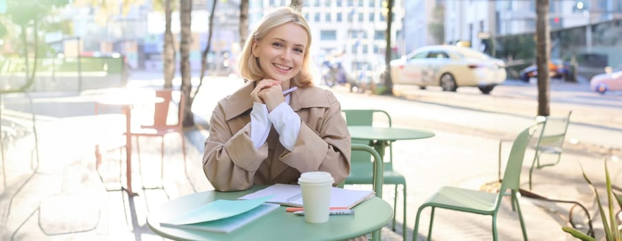 Portrait of beautiful young woman sitting in outdoor cafe with cup of coffee, reading through documents, student doing her homework on fresh air, smiling at camera.