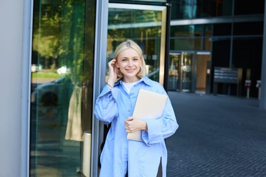 Lifestyle portrait of young woman with laptop, posing near campus, entrance to office building, holding backpack, smiling and looking happy.