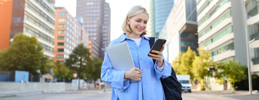Image of young stylish, modern blond woman in blue shirt, holding laptop and backpack, using smartphone, smiling, walking along the street, posing in city centre.