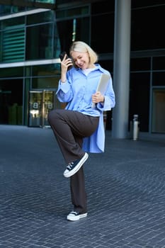 Vertical shot of stylish, happy modern blond girl, student dancing with smartphone near office building, saying yes, making fist pump, celebrating, feeling excitement.