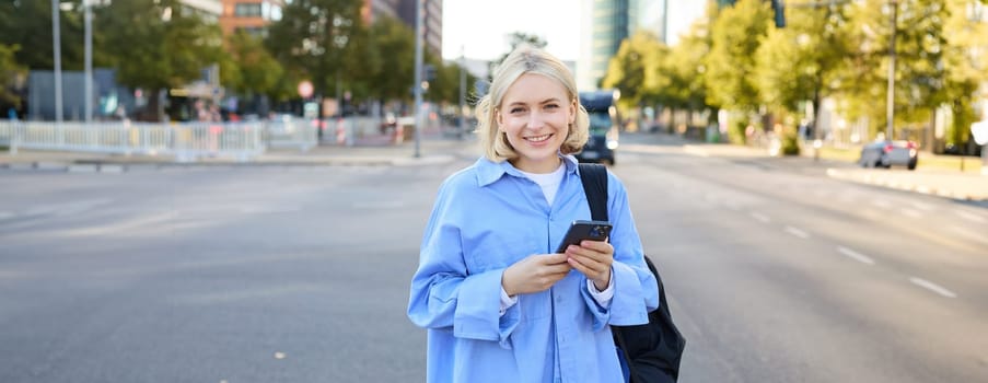 Portrait of blond smiling woman in blue shirt, holding backpack and smartphone, waiting for her ride, order taxi and checking updates on mobile phone, standing on street near road.
