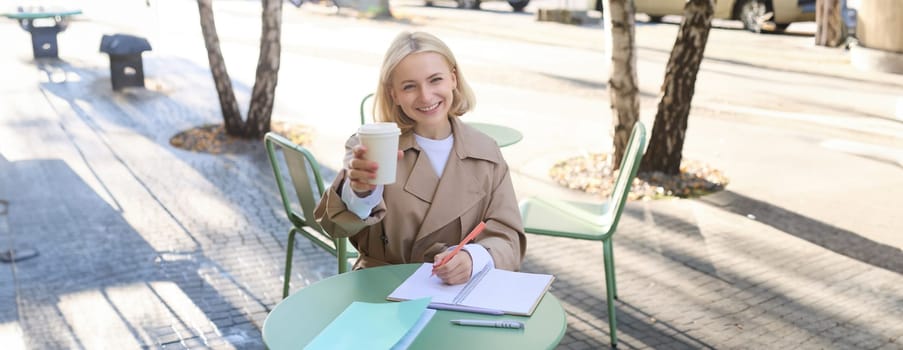 Beautiful, happy young blond girl working, writing in notebook, holding cup of coffee, drinking in cafe, sitting outdoors.