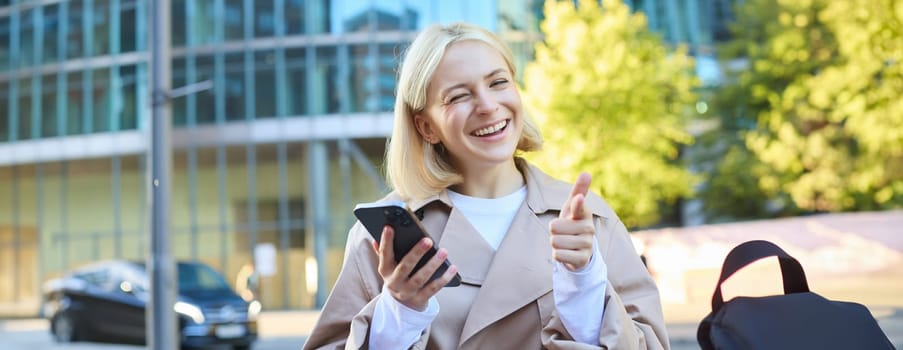 Cheerful young blond woman, university student, sitting on bench, showing thumbs up and winking at camera, holding mobile phone, recommending smth good.