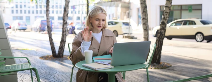 Image of excited blond girl, celebrating, sitting in coffee shop on sunny day, looking at laptop and triumphing. Lifestyle and emotions concept