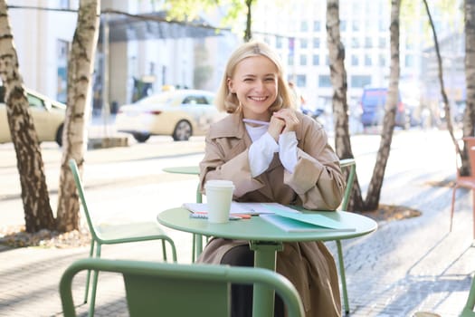 Portrait of young modern woman in stylish trench coat, sitting in outdoor cafe on sunny day, writing in her notebook, working or doing homework, drinking coffee.