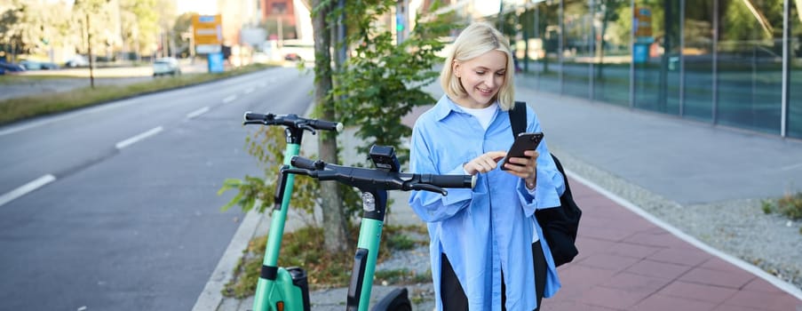 Image of smiling young woman heading to work, renting, using mobile phone app to unlock street scooter to get to her destination, posing on street with backpack.