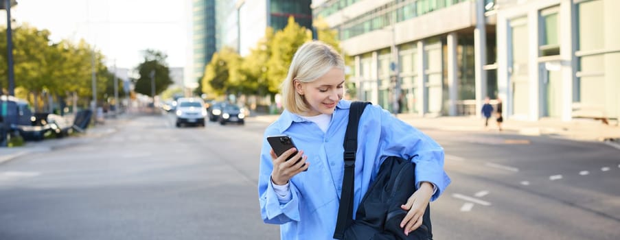 Lifestyle shot of young woman, student standing with smartphone on the street, opening her backpack pocket. People and occupation concept