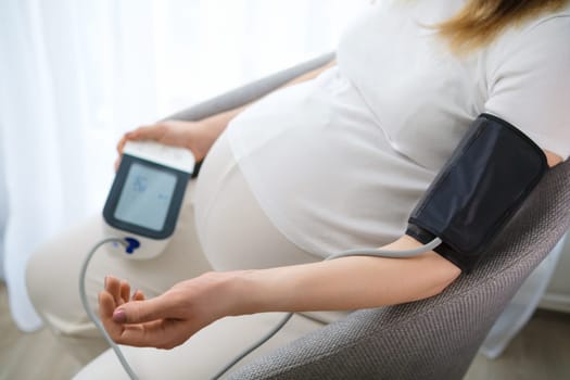 Future mother controls blood pressure with measuring instrument. Woman sits comfortably on chair looking at device expecting results crop body