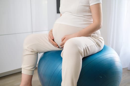 Pregnant woman slightly bouncing on rubber ball doing pregnancy gymnastics. Future mother gently touches growing belly in anticipation of baby crop body