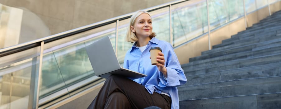 Lifestyle shot of young woman sitting on stairs with laptop, studying outdoors, drinking cup of coffee while working on computer, e-learning, studying on street.