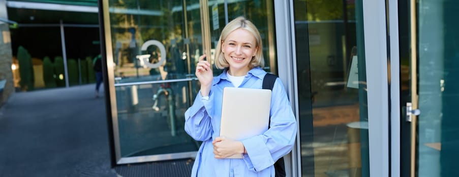 Portrait of carefree young woman, student with laptop, and backpack, leaning on campus building, smiling and laughing, studying and receiving scholarship in university.