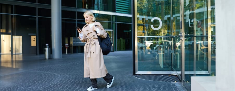 Portrait of blond young woman walking on street, student with backpack looking at her mobile phone, reading message on smartphone.