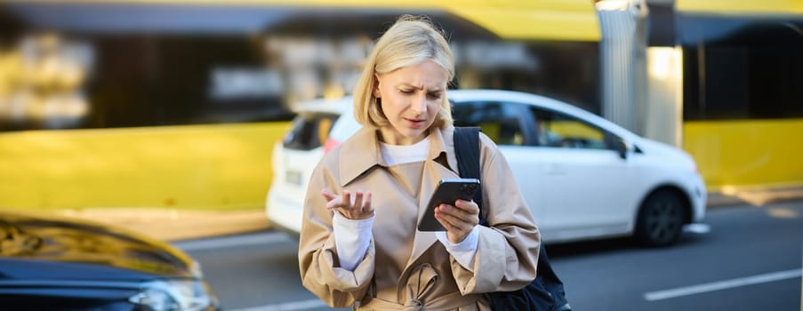 Portrait of young confused blonde woman, standing on busy street with cars behind her, looking at smartphone with puzzled face, shrugging shoulders, reading smth with clueless expression.