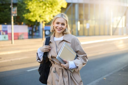 Portrait of young stylish female student, woman with backpack and notebook, smiling happy at camera, standing on street on sunny autumn day.