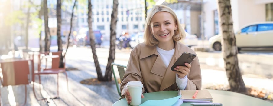 Portrait of young modern woman, student having her morning cup of coffee in city cafe, sitting outdoors, using mobile phone at the table, smiling at camera.