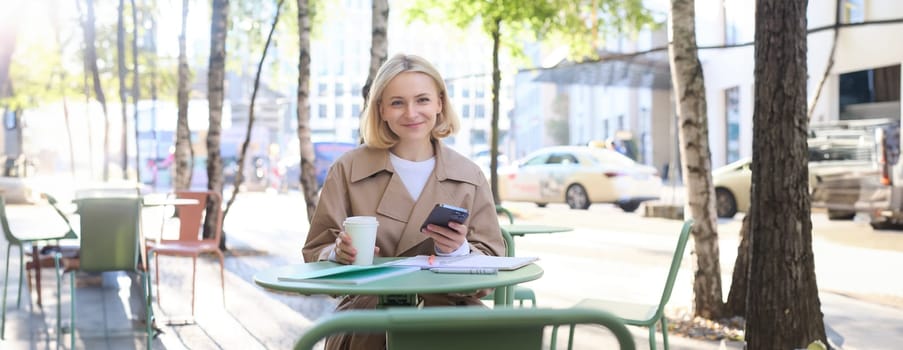 Portrait of young woman studying, sitting in outdoor cafe with smartphone and journals, doing homework, wearing trench coat on sunny day.