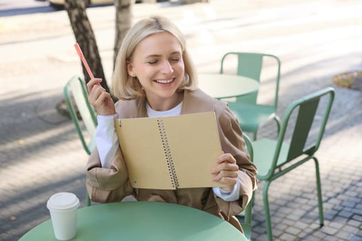 Portrait of laughing, smiling young woman, modern creative girl with notebook and pen, sitting in outdoor coffee shop and drawing, writing in journal, working on project.