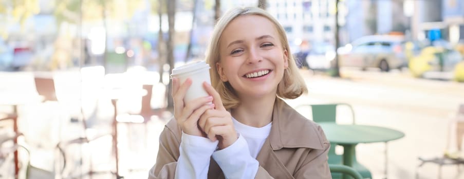 Close up portrait of laughing, smiling young woman, drinking cup of coffee and looking happy at camera, sitting in outdoor cafe on sunny bright day.