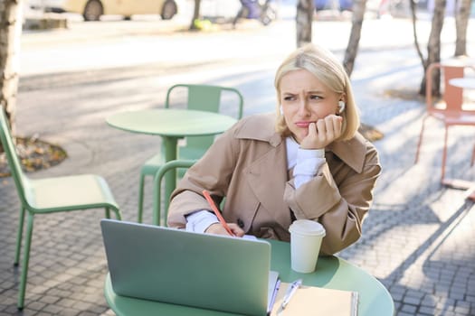 Portrait of young blond woman with laptop, sitting on street, drinking coffee in outdoor cafe, working, wearing wireless headphones, doing homework, freelancing.