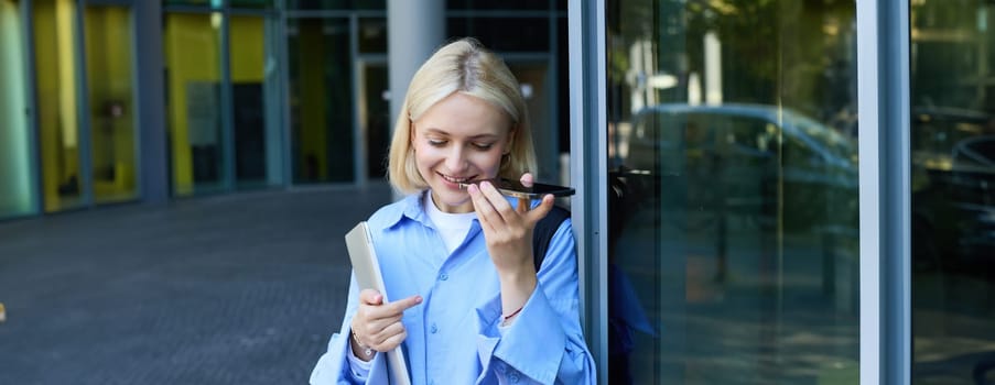 Portrait of smiling blond woman listening to message, recording voice on smartphone and looking happy, standing near office building with laptop.