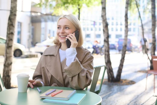 Close up of cheerful blond girl, woman talking on mobile phone, sitting in city centre, outdoor cafe, taking break from work or studying, chatting on telephone.