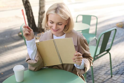Image of young smiling woman, cute girl writing in notebook, sitting outdoor cafe with pen and journal, drawing sketches outside on sunny day.