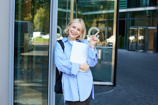 Portrait of smiling young woman, student or office worker with laptop, pointing finger at upper right corner, showing building and looking happy.