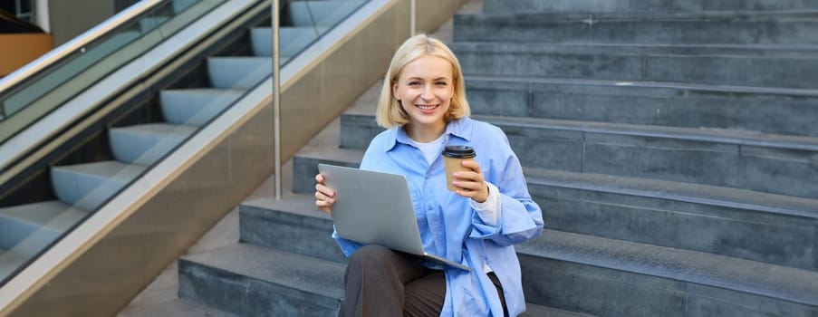 Young woman sitting with cup of coffee and laptop on stairs outdoors, working remotely, freelancing, connecting to public wifi in city to study online.