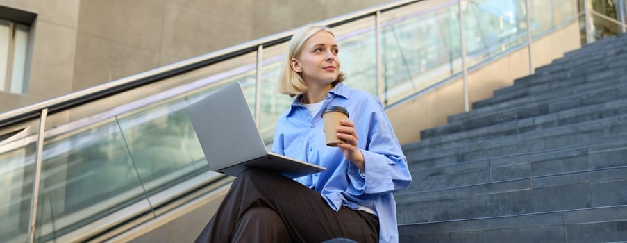 Image of stylish young woman, college student with laptop, sitting on city stairs and drinking coffee, working on project, studying outdoors, connects to public wifi, enjoying weather.