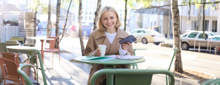 Portrait of modern young woman in city centre, sitting in cafe outdoors, drinking coffee, using mobile phone, smiling.