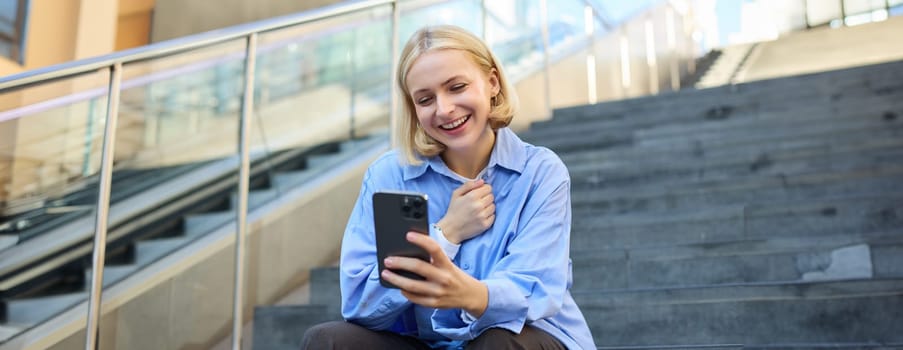 Image of young happy woman, sitting on stairs outside on street, taking selfie on smartphone camera, posing for photo social media profile, smiling.