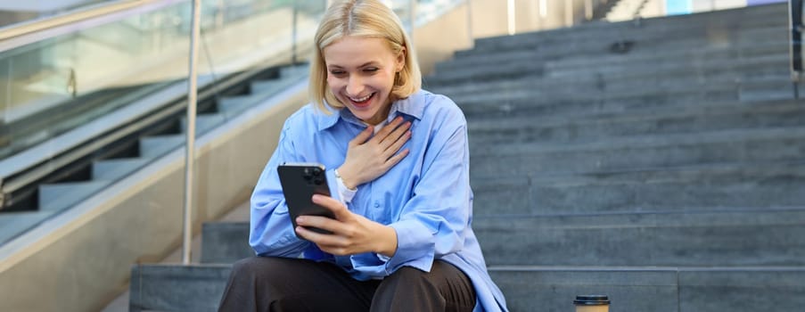 Portrait of young smiling woman, looking at smartphone with pleased, happy face, holding hand on chest, pleased to see something, sitting on stairs outdoors.