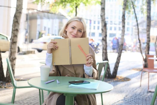 Outdoor shot of young beautiful blond girl with notebook, woman sitting in outdoor cafe, writing in journal, making plans in her planner, putting notes.