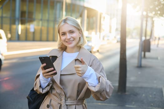 Lifestyle portrait of happy smiling woman, girl standing on street with smartphone, showing thumbs up with pleased face, recommending something good.