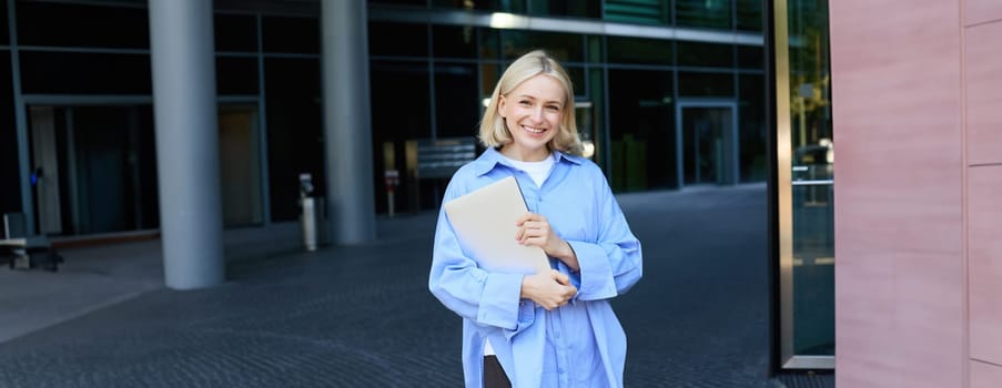 Portrait of young blond woman, student standing near her campus with notebooks and documents, wearing blue shirt and smiling at camera. Education concept