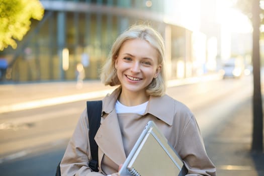 Image of beautiful young smiling woman with blond hair, walking along street on sunny day, carry her notebooks and college materials, looking carefree.