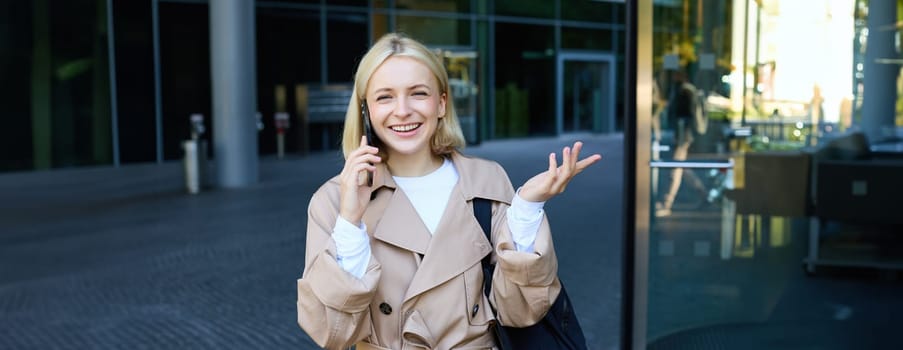 Carefree young woman with smartphone, walking outdoors in city centre, standing on street and chatting, talking on mobile phone with happy, cheerful smiling face expression.