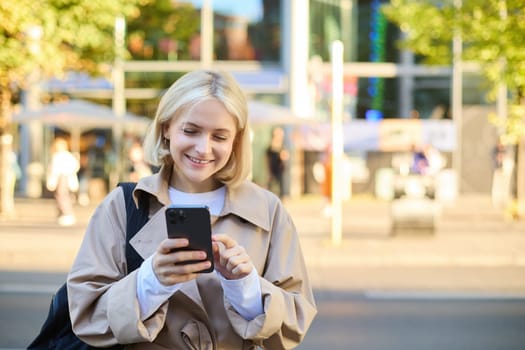 Close up portrait of young smiling blond woman, student on street, using mobile phone, checking messages on smartphone, walking on road.