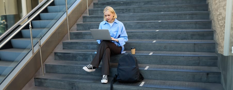 Freelance employee, woman working on her project, using laptop, sitting outdoors on stairs, drinking takeaway coffee.