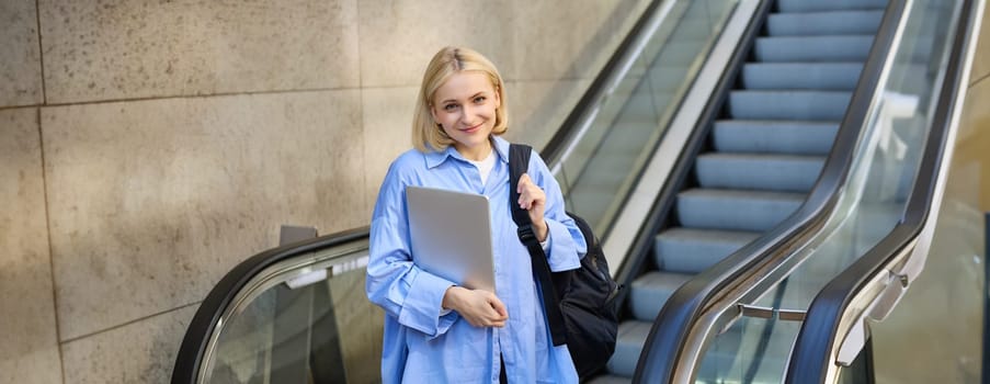 Education and people concept. Young woman with laptop and backpack, using escalator in city centre, smiling and looking at camera.