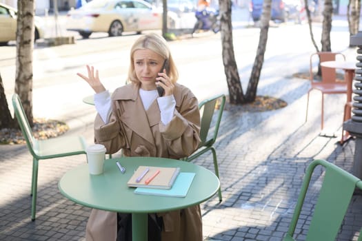 Portrait of young woman sitting alone in cafe, outdoors table, looking upset and disappointed, answer phone call, talking on smartphone and shrugging.