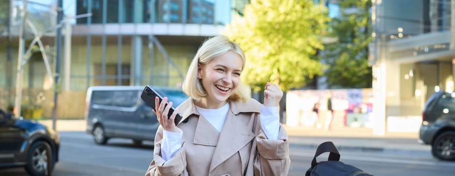 Outdoor shot of cheerful young woman with smartphone in hand, sitting on street bench and rejoicing, making fist pump, celebrating victory, winning smth.