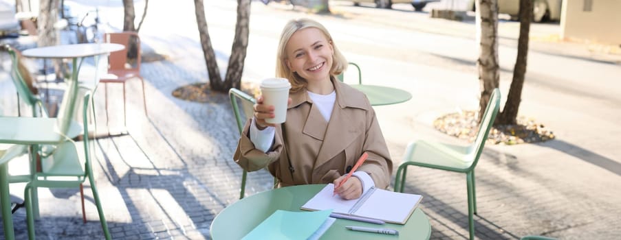 Smiling young blond woman, enjoying her coffee, drinking takeaway and sitting in street cafe, working on project, student doing homework, writing something in notebook, looking happy at camera.