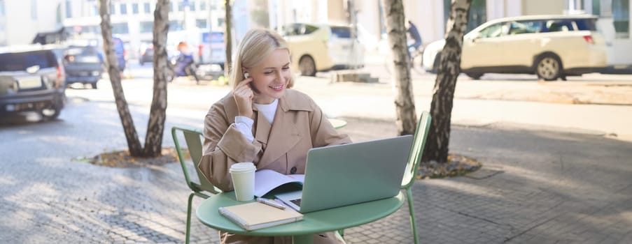 Portrait of young stylish woman, student in outdoor coffee shop, using laptop and making notes, wearing wireless headphones, listening to online course.